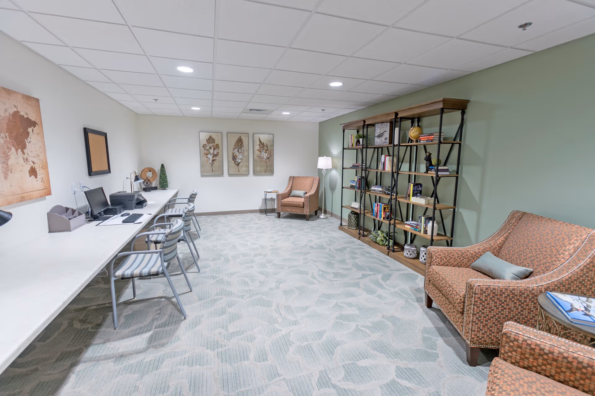 A well-lit room with a long white desk along one wall featuring three chairs, a computer, and a printer. On the opposite wall, there is a large bookshelf filled with books and decorative items. Two patterned armchairs with a small round table between them are positioned near the bookshelf. The room has light green and white walls, a patterned carpet, and ceiling lights.