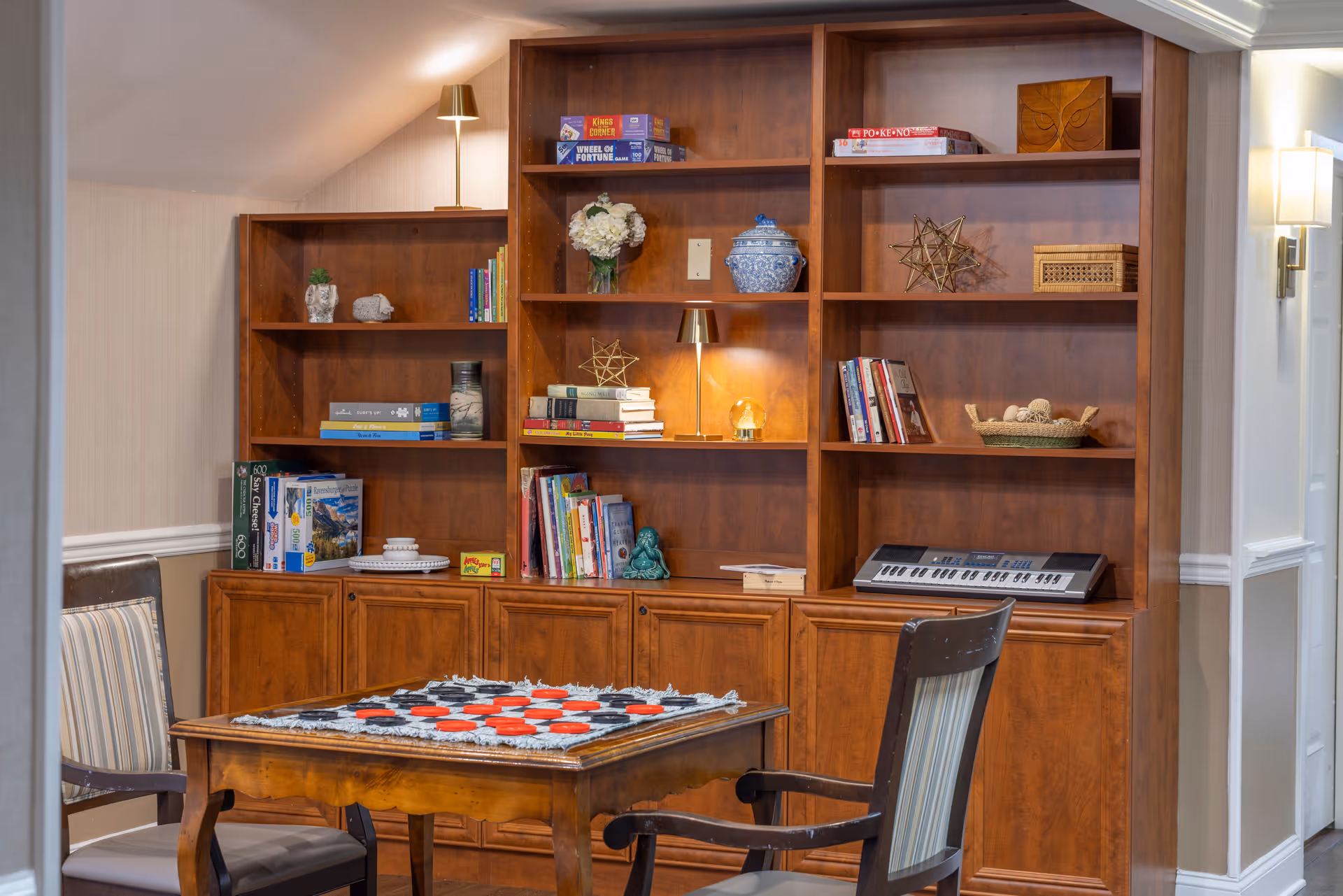 A cozy interior room with a wooden bookshelf filled with books, board games, decorative items, and a small electronic keyboard. In front of the bookshelf is a wooden table with a checkers game set up, surrounded by two chairs with striped cushions. The room has warm lighting and beige walls.