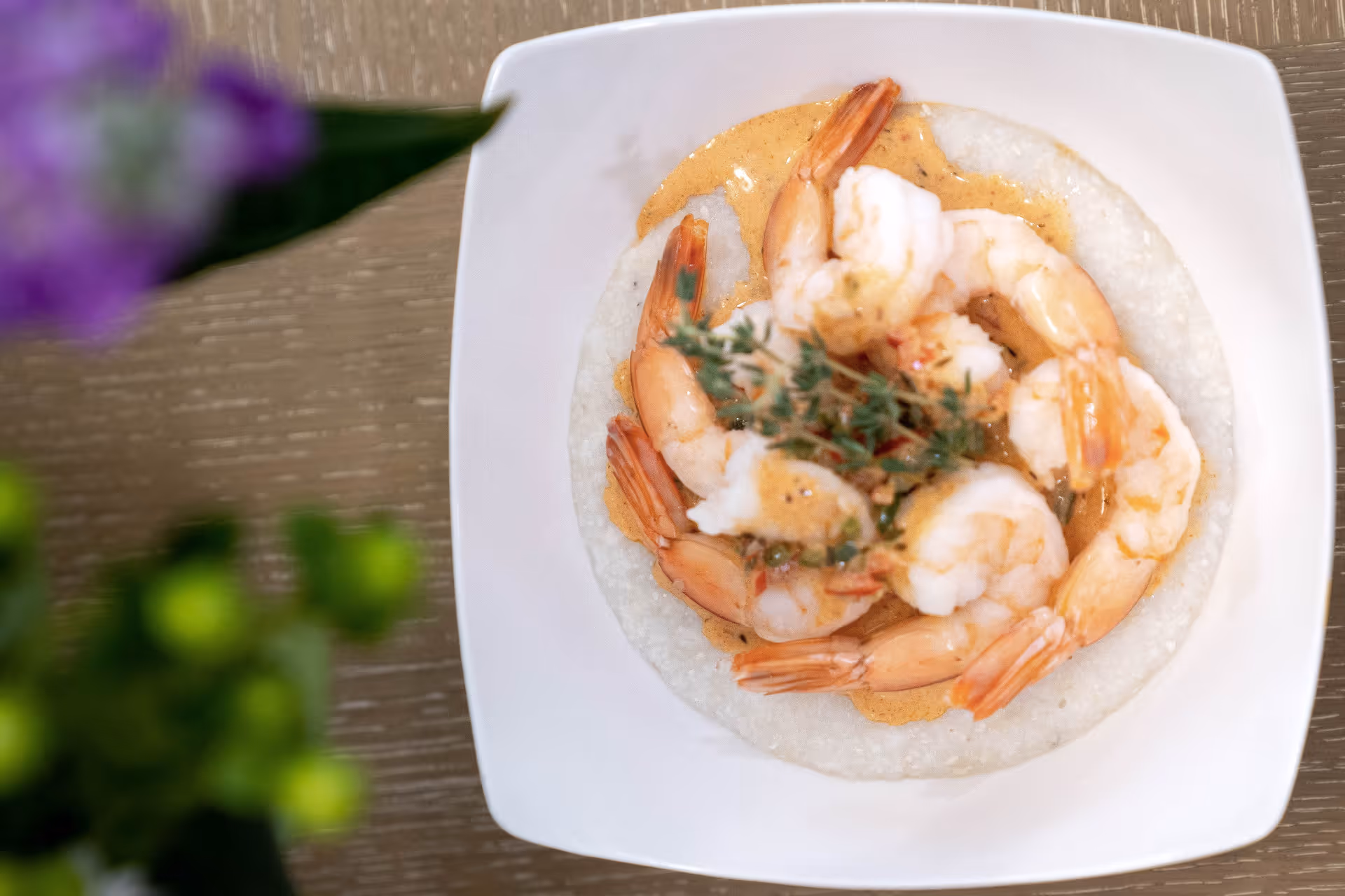 A white square plate with a serving of shrimp arranged in a circle on a bed of creamy grits, garnished with a sprig of fresh herbs, on a wooden table with blurred green and purple plants in the foreground.