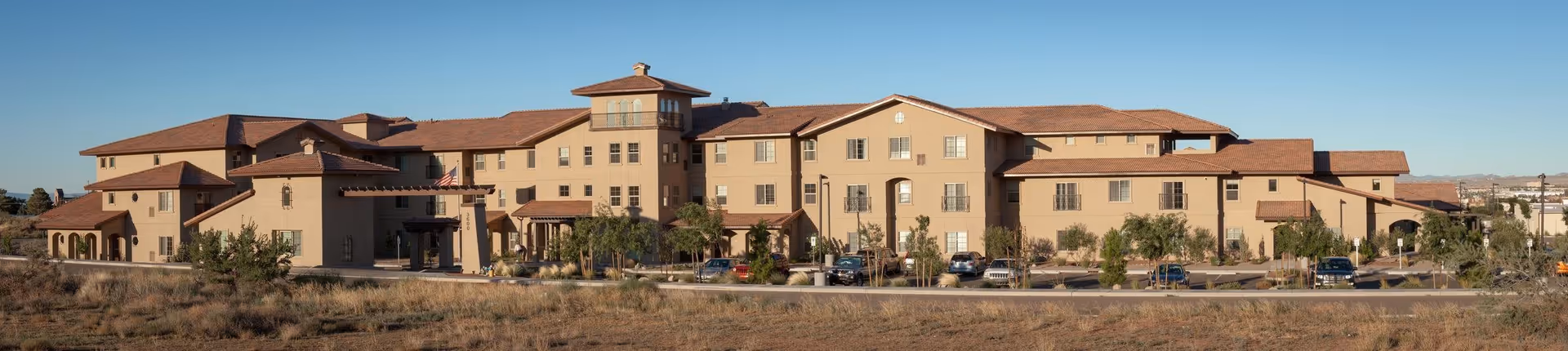Wide exterior view of White Cliffs Senior Living facility, a large multi-story building with beige walls and a red-tiled roof, surrounded by a parking lot with several cars and some landscaping including trees and shrubs under a clear blue sky.