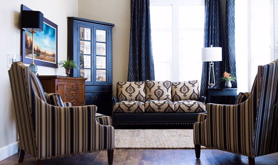 A cozy living room with a patterned beige and black sofa centered against a window with dark blue curtains. Two striped armchairs face the sofa. There is a wooden cabinet with drawers and a black display cabinet with glass doors on the left side. A side table with a lamp and a small plant is on the right side of the sofa. The floor is wooden with a light-colored rug in the center.