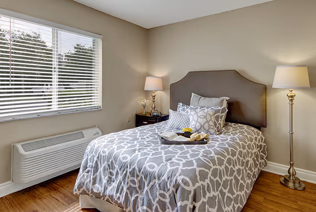 A neatly made bed with a gray and white patterned comforter and multiple pillows in a bedroom. There is a tray with a book, a cup, and a small flower on the bed. The room has wooden flooring, a window with blinds, a dark wooden nightstand with a lamp and flowers, and a tall floor lamp beside the bed.