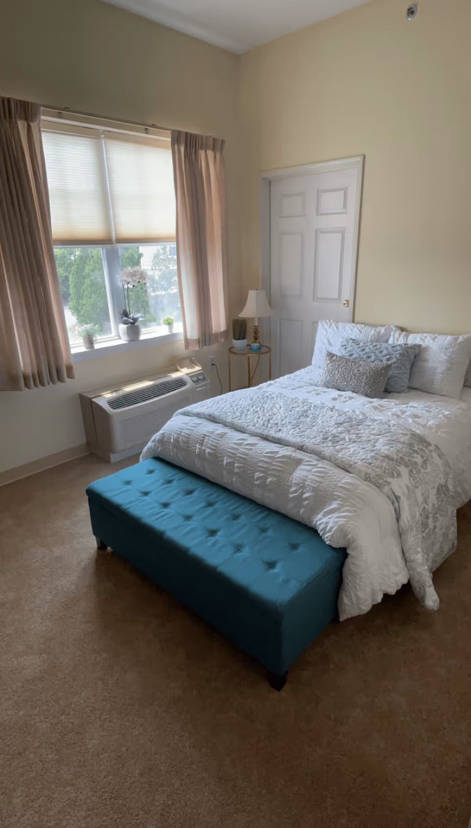 A neatly made bed with white and gray bedding and decorative pillows in a bedroom. At the foot of the bed is a teal tufted bench. To the right of the bed is a small round side table with a lamp and decorative items. A window with beige curtains and a white blind lets in natural light. Below the window is a wall-mounted air conditioning unit. The walls are painted light beige, and there is a closed white door in the corner.