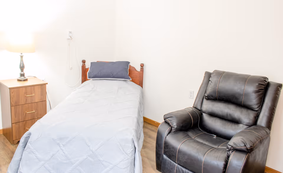 Small bedroom with a single bed, wooden nightstand and lamp, and a black recliner against a white wall.