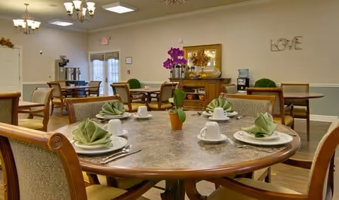 A dining room in a senior living facility with round tables set with white plates, cups, and green folded napkins. There is a purple orchid centerpiece on the nearest table. The room has beige walls, chandeliers, and a sideboard with decorative items including a framed picture and a 'LOVE' wall decoration.