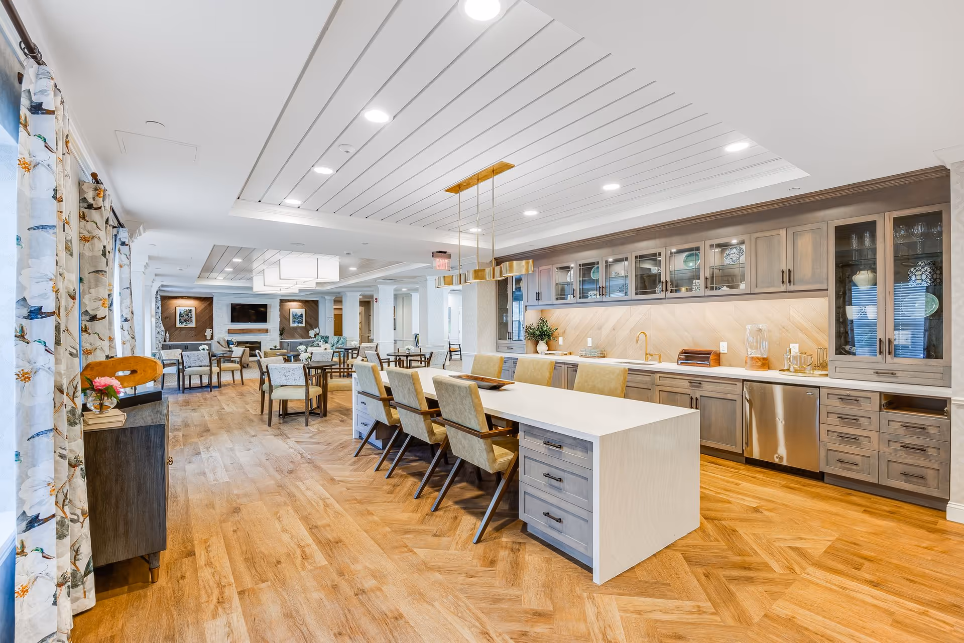 A spacious and well-lit common area in a senior living facility featuring a modern kitchen with wooden cabinets, a long white island with six beige chairs, and a dining area with multiple tables and chairs. The room has wooden flooring, floral curtains on the left, and a cozy seating area with a fireplace and TV in the background.