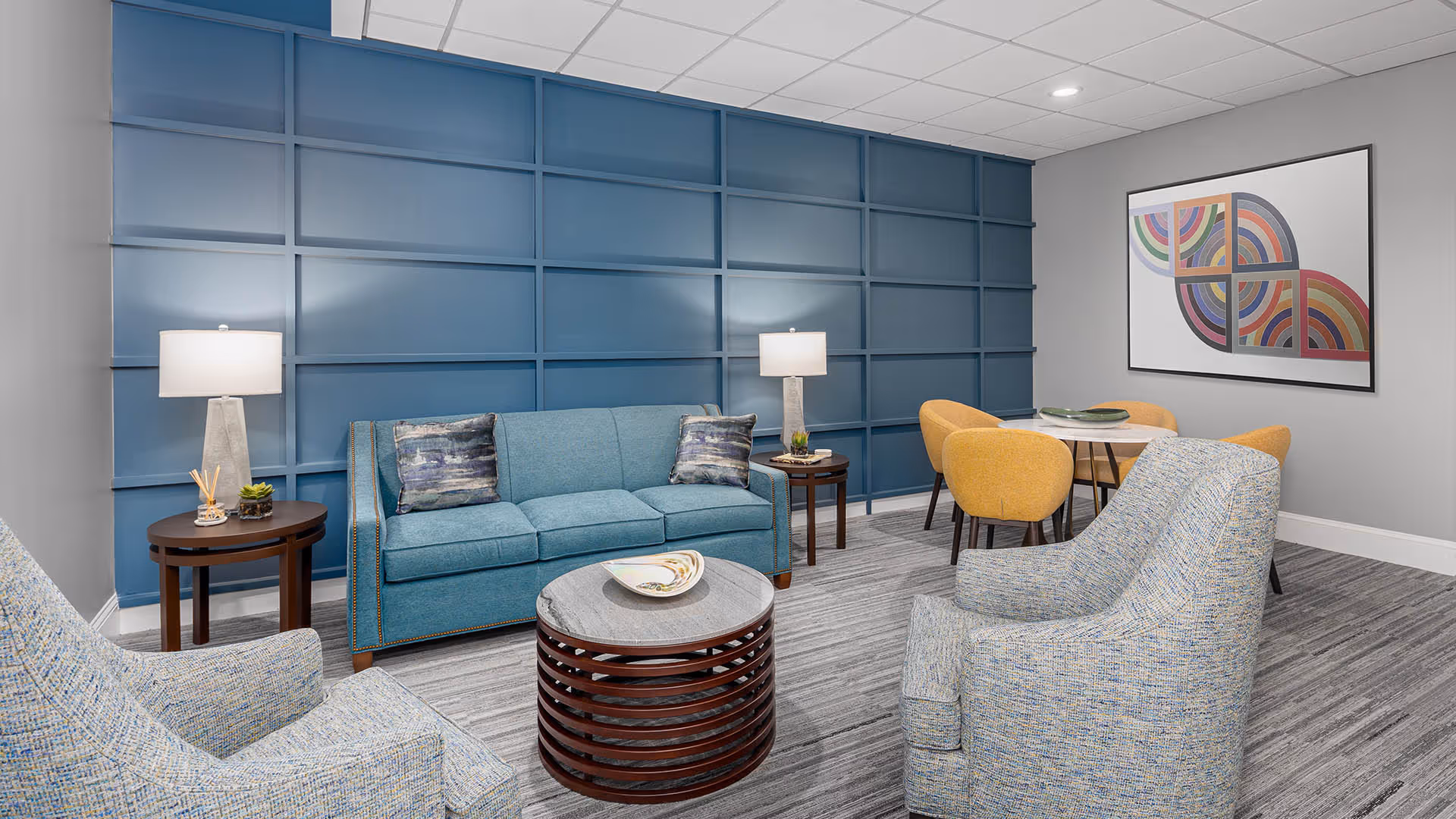 A modern living room with a blue accent wall featuring a grid pattern. The room includes a blue sofa with two patterned pillows, two light blue armchairs, a round coffee table with a decorative bowl, and two side tables with lamps and small plants. In the background, there is a small dining area with a round white table and four yellow chairs, and a colorful abstract artwork on the wall.