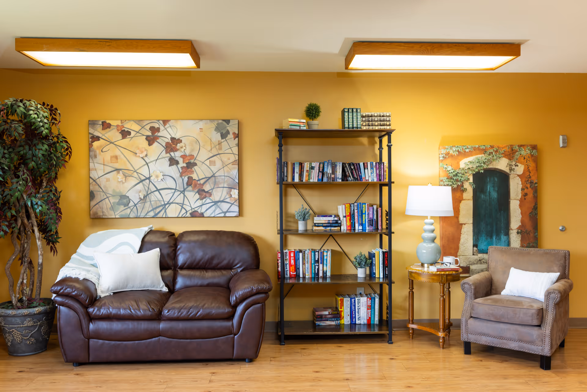Cozy seating area with a brown leather loveseat, armchair, bookshelf, lamp, and wall art against a warm yellow wall.