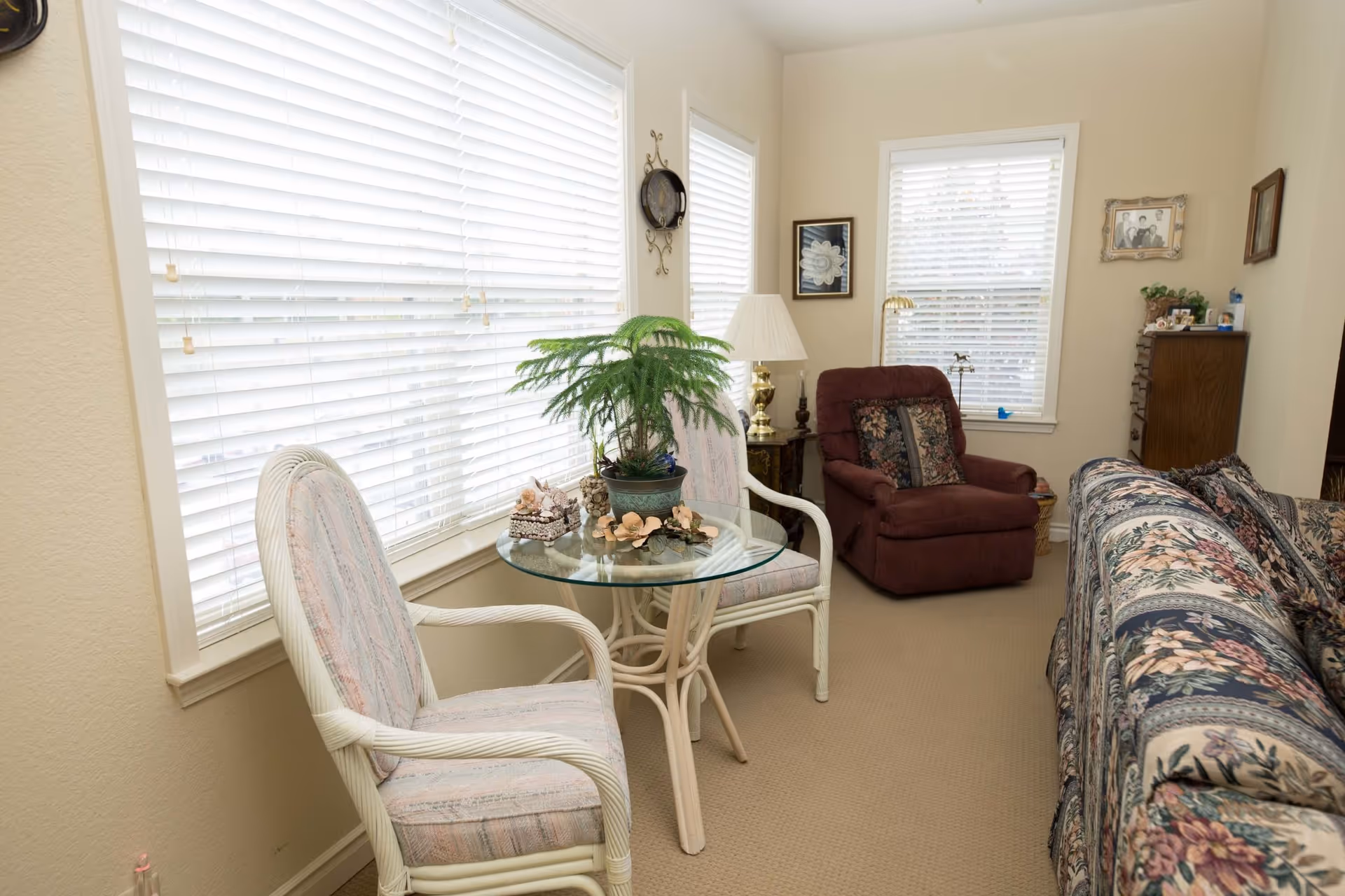A cozy living room area with large windows covered by white blinds. There are two cushioned wicker chairs with a glass-top table between them, decorated with a potted plant and seashell ornaments. A floral-patterned sofa and a maroon recliner chair with a floral cushion are also visible. The walls are adorned with framed pictures and a brass lamp sits on a side table.