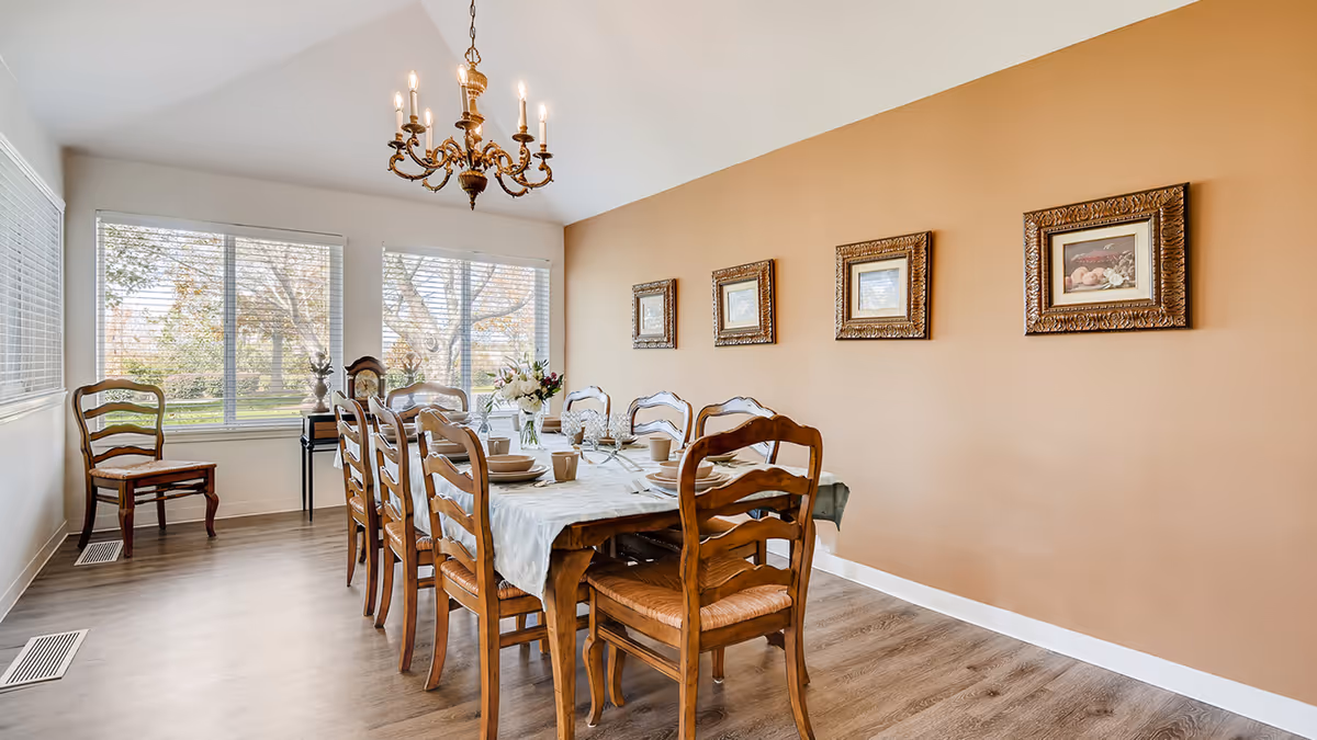 Bright dining room with a long wooden table set for a meal, chandelier overhead, large windows, and framed artwork on a tan wall.