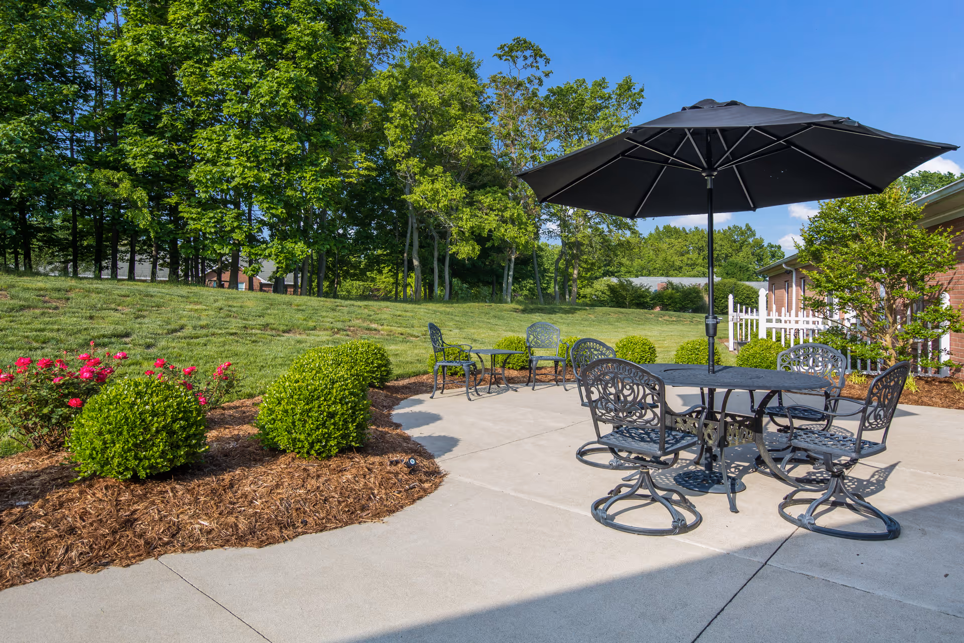 Outdoor patio area with black metal table and chairs under a large black umbrella, surrounded by green bushes and trees with a clear blue sky.