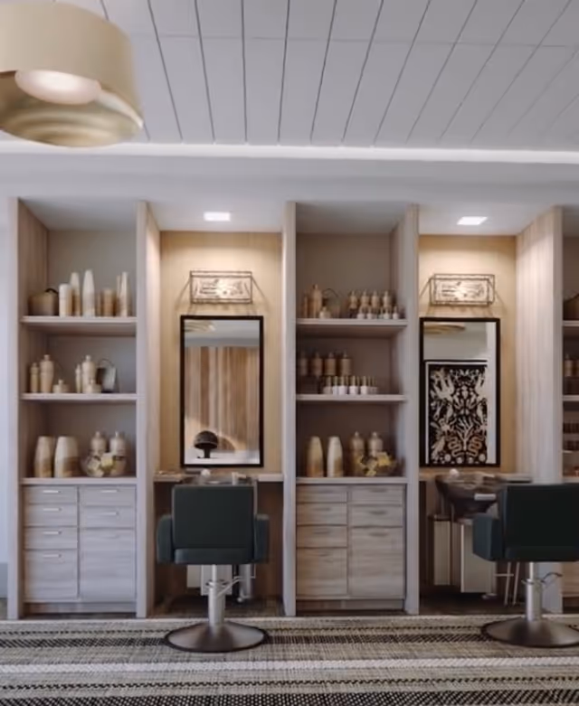 Interior view of a salon area with two styling stations. Each station has a large mirror, a black salon chair, and shelves filled with various bottles and containers. The space features light wood cabinetry and a patterned rug on the floor.
