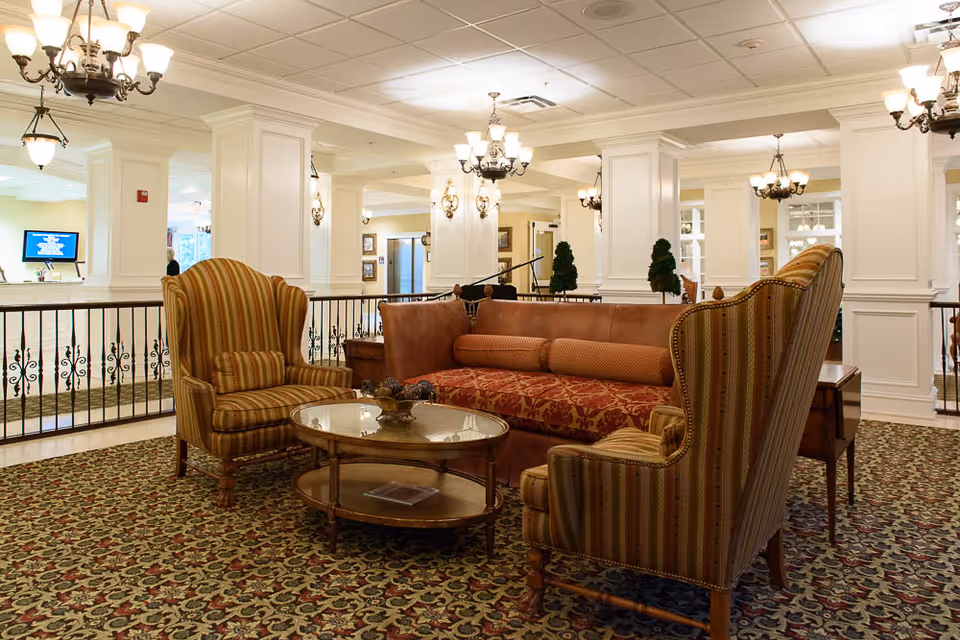 A cozy seating area in a senior living facility featuring a patterned carpet, a brown sofa with decorative cushions, two striped armchairs, and a round glass coffee table. The room has white paneled columns, elegant chandeliers, and wall sconces providing warm lighting. In the background, there is a reception desk with a computer monitor and a person standing behind it.