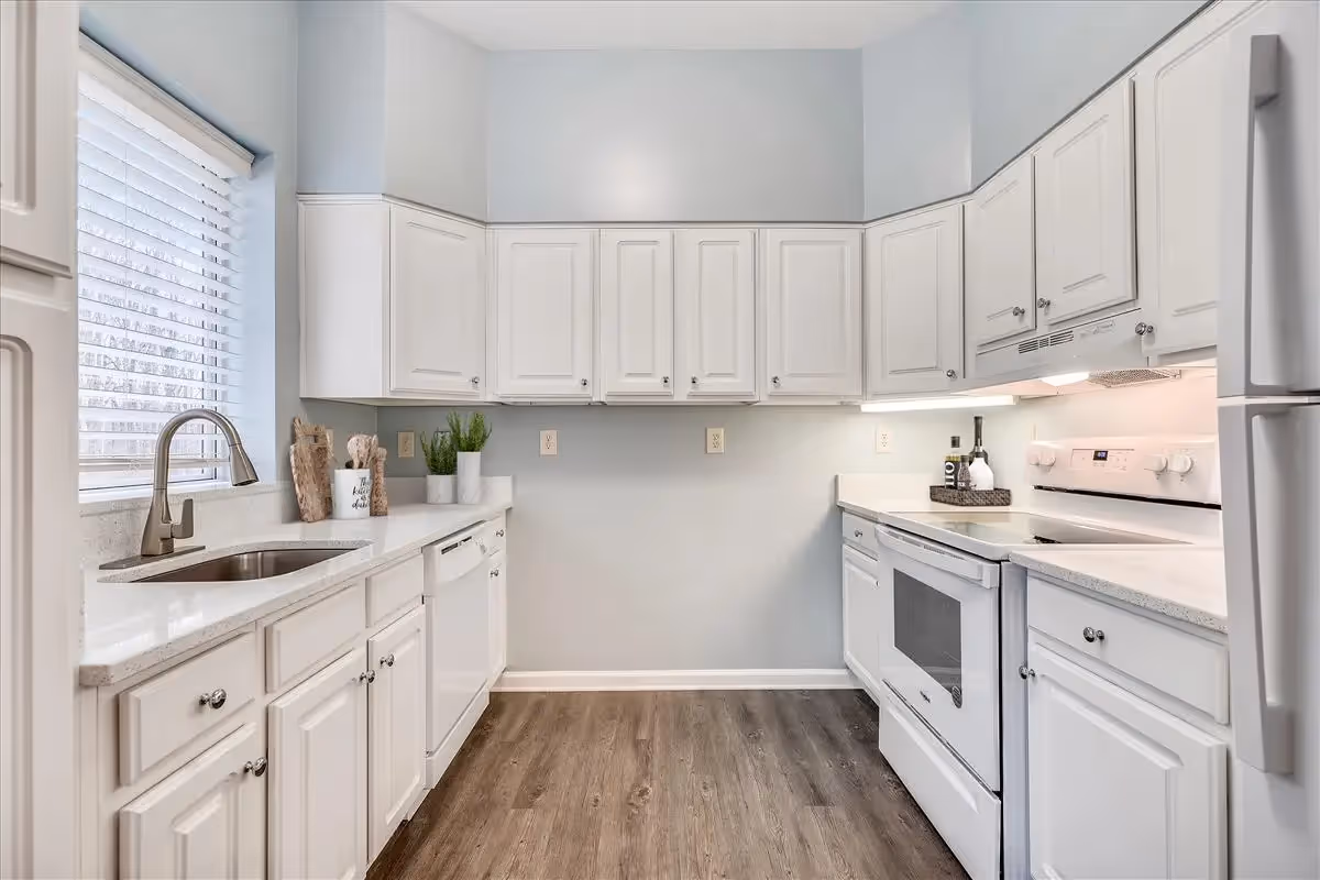 A bright and clean kitchen with white cabinets, white countertops, a stainless steel sink under a window with blinds, a white dishwasher, a white stove with an oven, and a white refrigerator. The floor is wood-style laminate, and there are small decorative plants and kitchen utensils on the countertops.