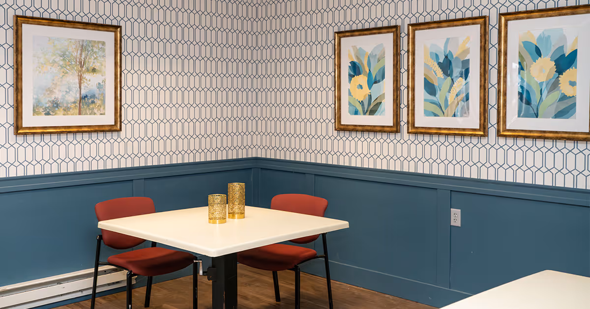 A small corner dining area with a square white table and two red chairs. The walls have a geometric patterned wallpaper above blue wainscoting. Four framed floral and tree artworks hang on the walls. Two gold decorative candle holders are on the table.