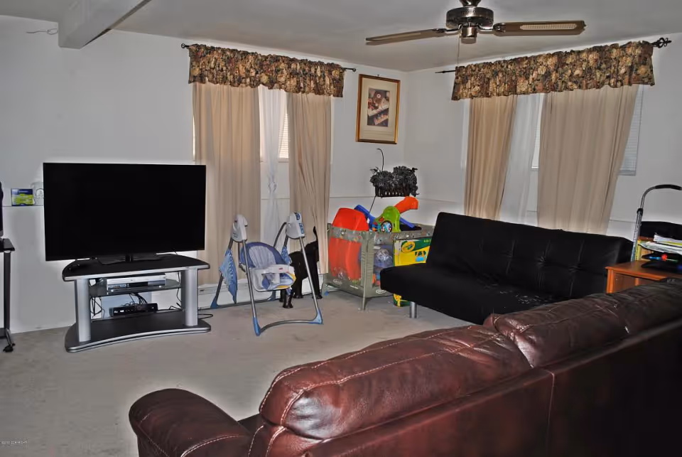 Living room with leather sofas, a flat-screen TV on a stand, a baby swing and playpen, and curtains over the windows.