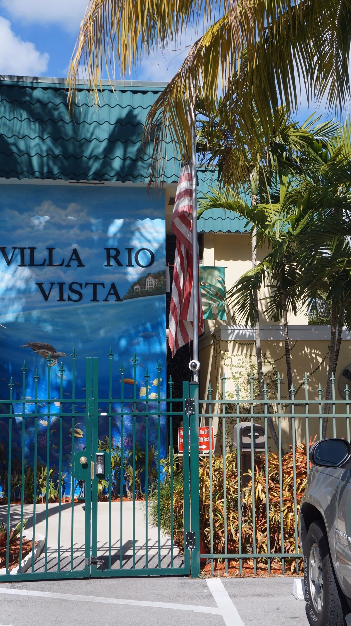Entrance gate of Villa Rio Vista facility with a green metal fence and gate, an American flag on a pole, tropical palm trees, and a mural on the building wall depicting an underwater scene with fish and a turtle. A silver vehicle is partially visible on the right side.