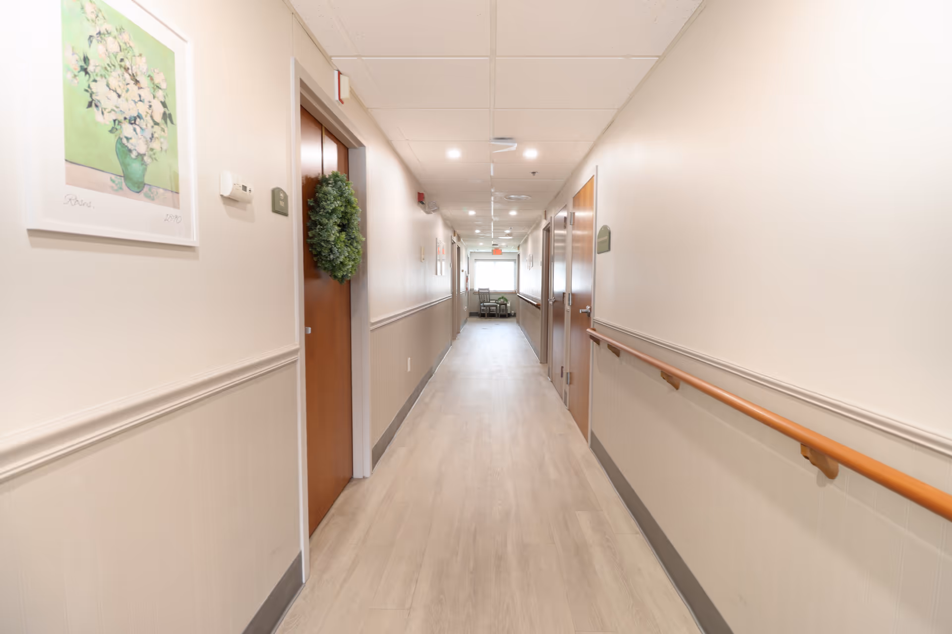 A long, well-lit hallway in a senior living facility with light-colored walls and wood flooring. The hallway has handrails on both sides and several closed wooden doors, one decorated with a green wreath. A framed picture of flowers hangs on the left wall, and at the end of the hallway, there are two chairs near a window letting in natural light.