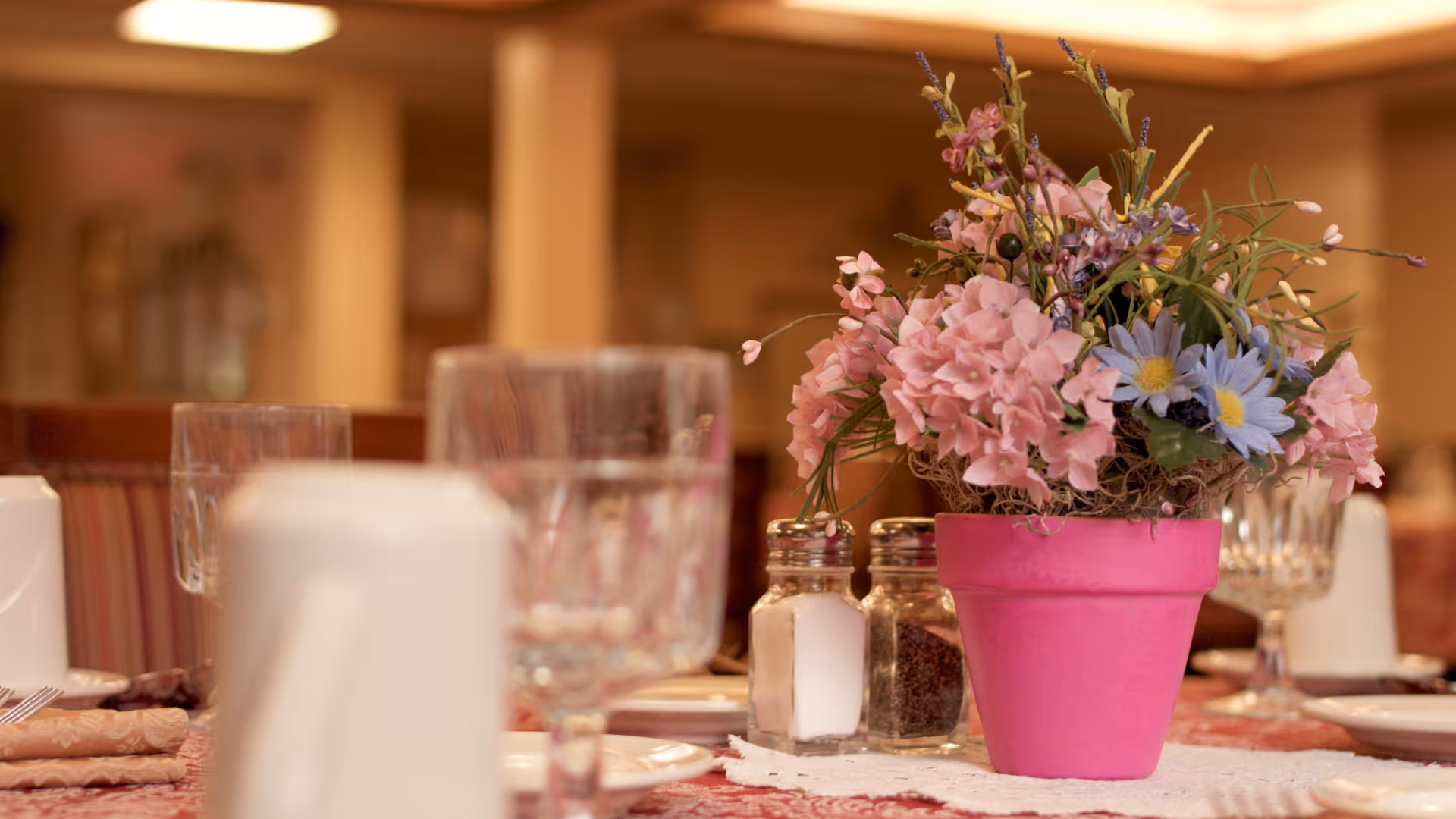 Table setting in a dining room with a pink potted flower centerpiece, salt and pepper shakers, and glassware.