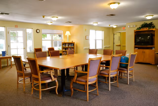A well-lit common room with a large rectangular wooden table surrounded by wooden chairs with purple cushions. The room has beige walls, carpeted floor, windows letting in natural light, a bookshelf with books, a lamp, and a TV cabinet with a television. There are exit doors and an exercise ball visible in the room.