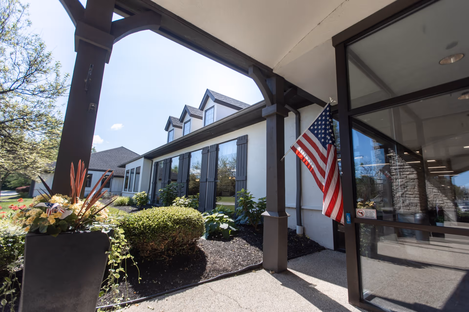 Entrance area of a senior living facility with a covered porch supported by dark wooden beams, an American flag mounted on one beam, large windows with black shutters, and well-maintained landscaping including bushes and flowers in a planter.