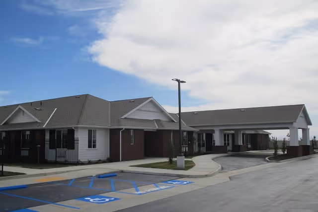 Exterior view of a single-story senior living facility building with a covered entrance driveway, several windows, and a parking lot with designated handicapped parking spaces under a partly cloudy sky.