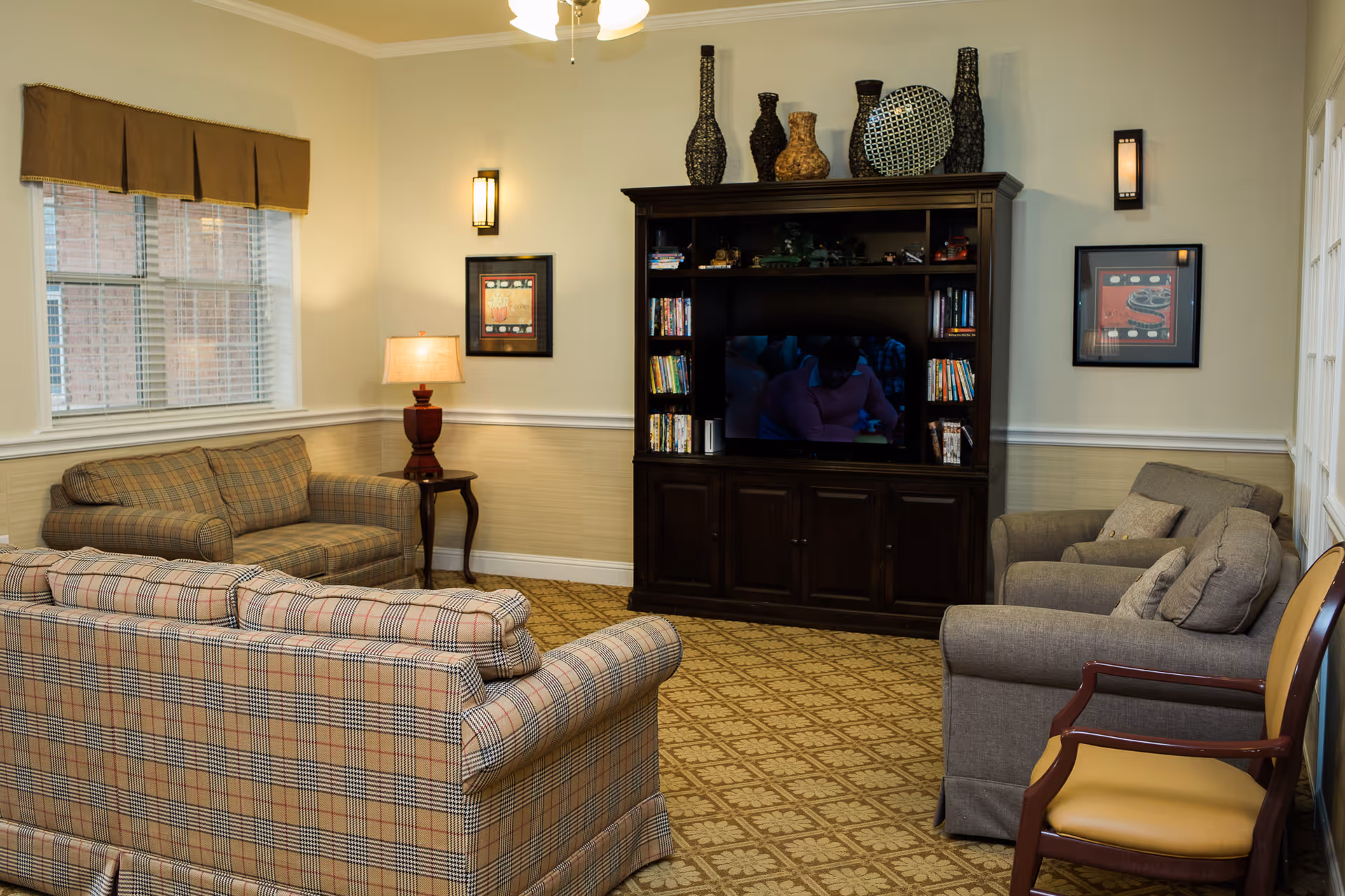 A cozy living room with patterned carpet, two plaid sofas, a gray armchair, a wooden chair with a yellow cushion, and a dark wooden entertainment center holding a TV, books, and decorative vases. The room has beige walls with two framed pictures, a window with brown valance, a table lamp on a small side table, and wall sconces providing warm lighting.