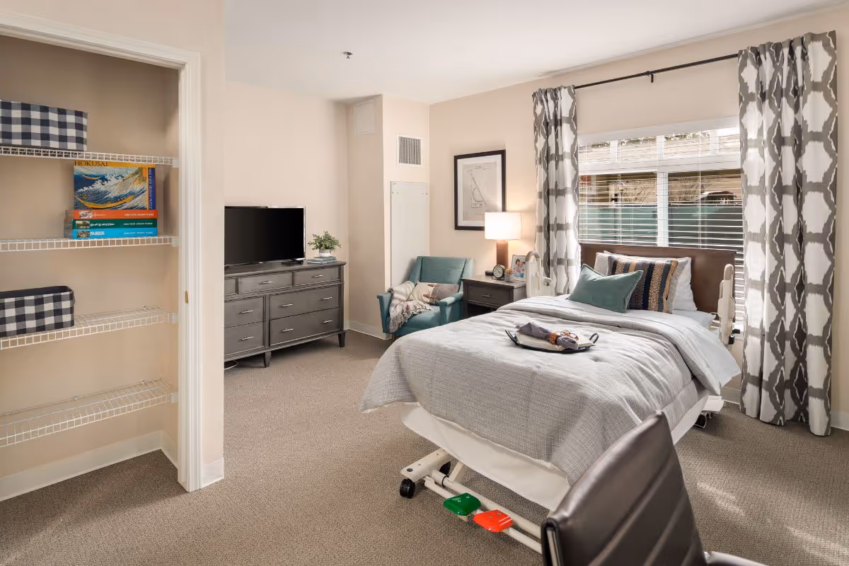 Furnished senior bedroom with an adjustable hospital-style bed, bedside table and lamp, armchair, dresser with TV, open shelving, and a window with patterned curtains.