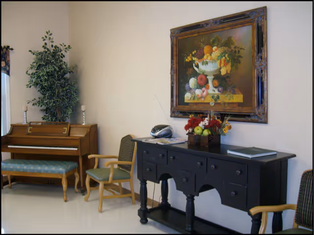 A cozy interior corner featuring a wooden piano with a cushioned bench, two armchairs with green cushions, a black sideboard with drawers, a decorative fruit and flower arrangement, a small radio, and a large framed painting of a fruit still life hanging on the wall.