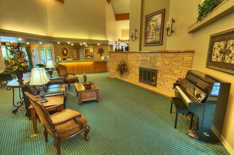 A spacious and warmly lit common area with green patterned carpet, featuring a black upright piano on the right, a stone fireplace with a framed painting above it, and several wooden chairs and tables with lamps and floral arrangements. In the background, there is a reception desk with a lit counter and decorative items, and large windows letting in natural light.