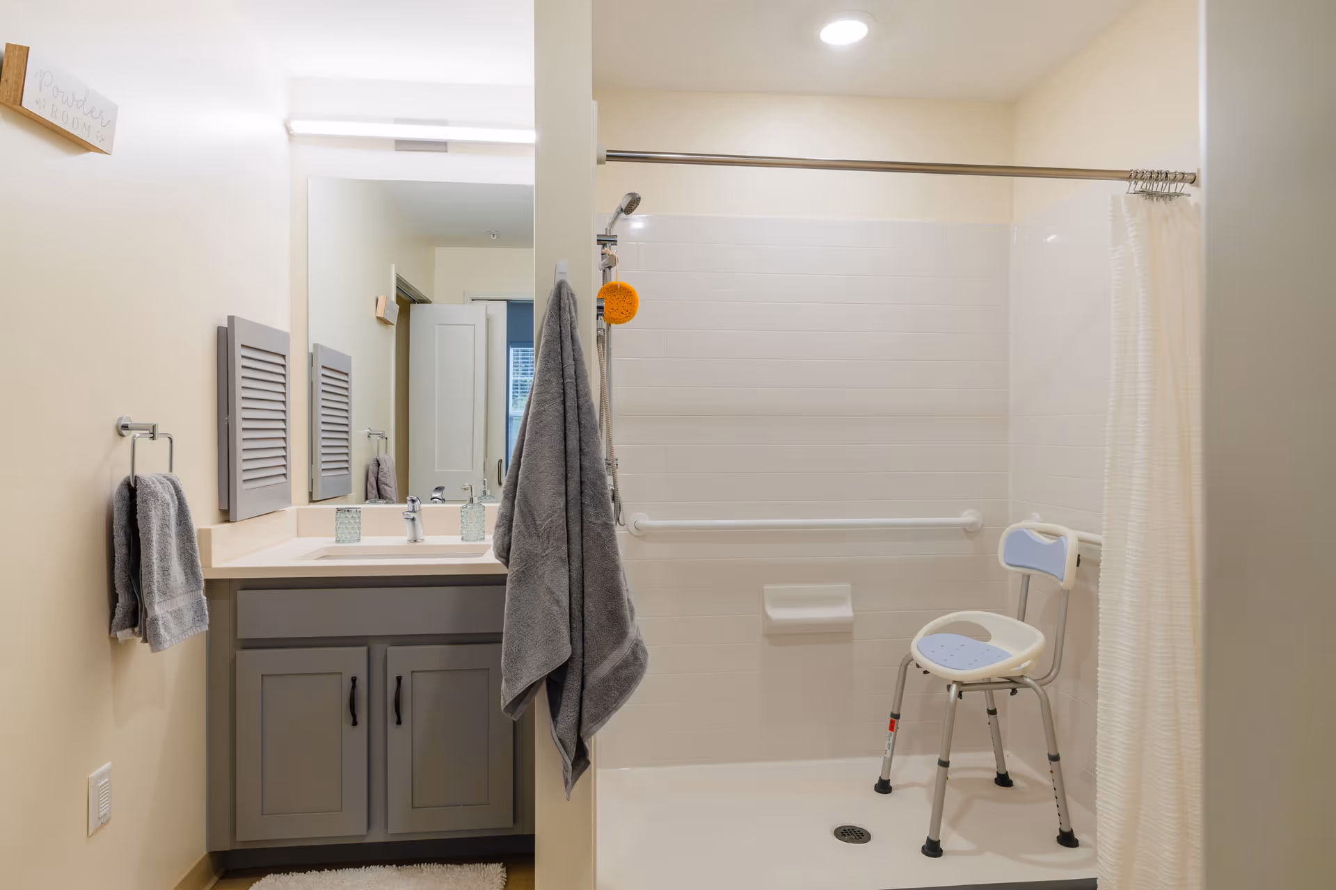 A clean and accessible bathroom with a gray vanity cabinet, a large mirror, and a sink. There are gray towels hanging on the wall and on a hook. The shower area has white tiled walls, a grab bar, a shower chair, and a white shower curtain. A small sign on the wall reads 'Powder Room'.
