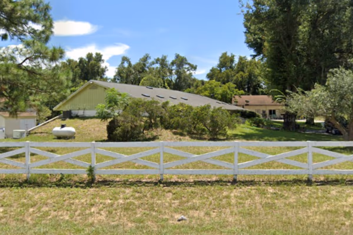 A single-story building with a sloped roof and several skylights, surrounded by trees and bushes. In front of the building is a white wooden fence and a grassy area under a clear blue sky.