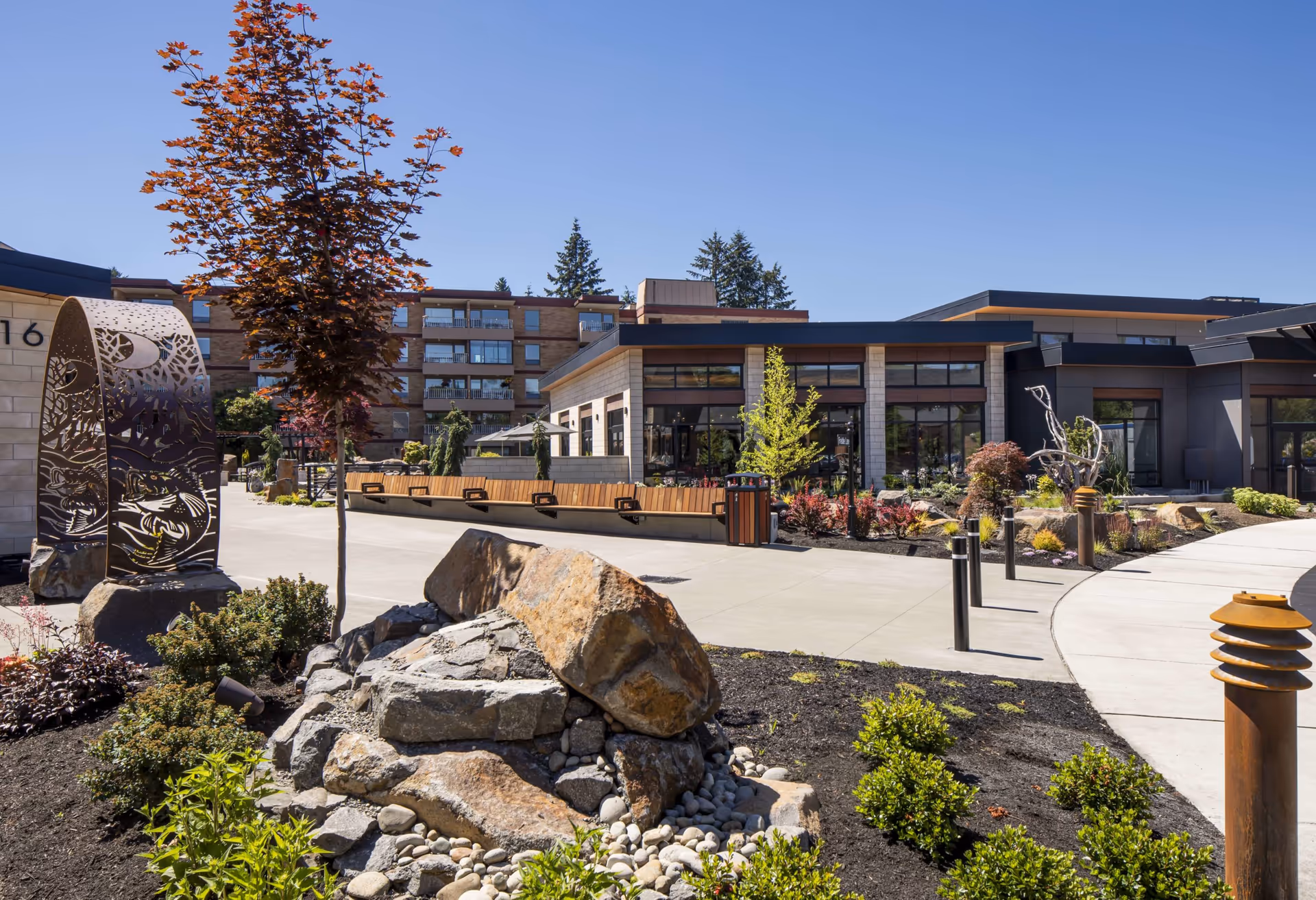 Outdoor view of a senior living facility named Panorama featuring landscaped gardens with rocks, shrubs, and trees, a paved walkway, modern building structures with large windows, and decorative metal sculptures under a clear blue sky.