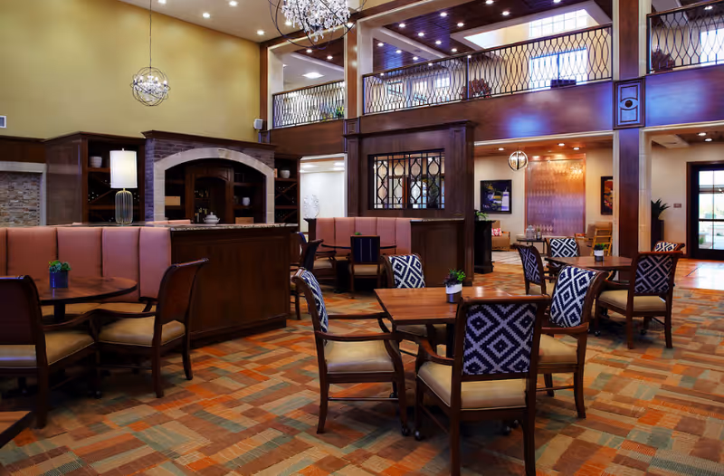 Spacious and elegant common area with multiple wooden tables and chairs, some with patterned upholstery, arranged on a carpeted floor. The room features warm lighting, a high ceiling with a chandelier, wooden beams, and a second-floor balcony with decorative railing. There are built-in shelves and a cozy seating area with cushioned benches.