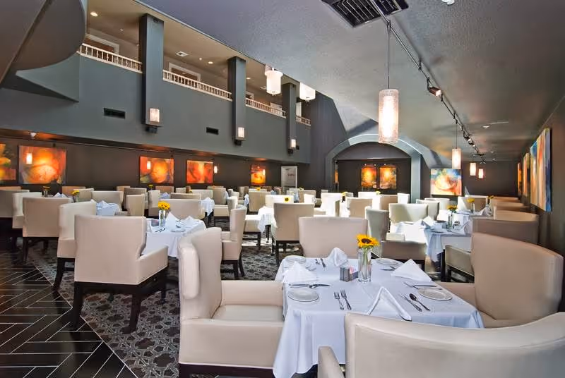 A spacious dining room with multiple tables covered in white tablecloths, each set with plates, silverware, napkins, and small vases holding yellow flowers. The room features beige upholstered chairs and booths, dark walls adorned with colorful abstract paintings, and modern pendant lighting hanging from a curved ceiling.