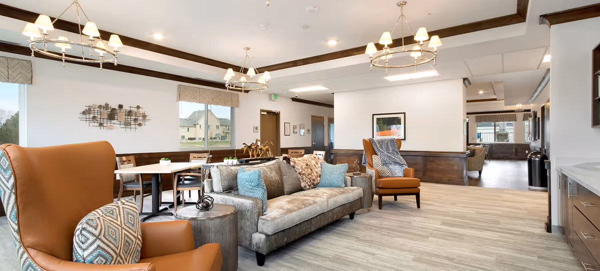 A bright and spacious senior living common area with comfortable seating including a gray sofa with blue and patterned pillows, two brown armchairs with patterned cushions, and small round side tables. The room features large windows with views of neighboring buildings, modern chandeliers, and light wood flooring. The walls are white with dark wood trim and decorative wall art.