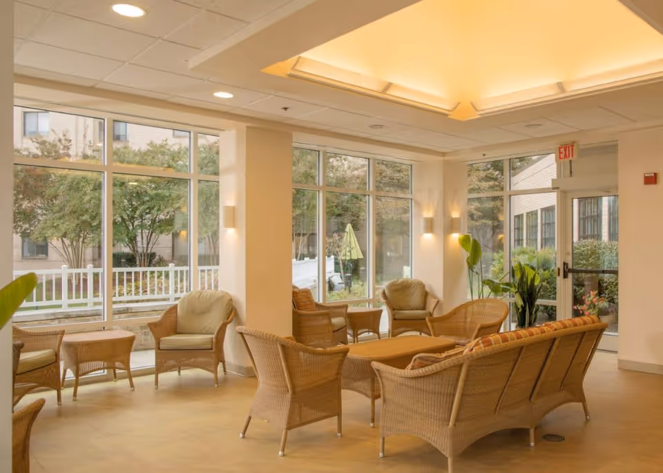 A bright and spacious common area with large windows showing an outdoor garden. The room is furnished with wicker chairs and a sofa with cushions arranged around a central coffee table. There are plants near the windows and a door with an exit sign above it.