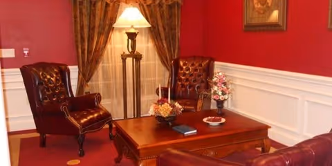 A cozy living room with red walls and white wainscoting, featuring two dark brown leather armchairs, a wooden coffee table with floral arrangements and a book, a floor lamp with a beige shade, and heavy patterned curtains covering a window.