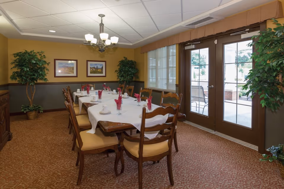 A dining room with a long set table, wooden chairs, potted plants, framed artwork, and glass doors opening to an outdoor area.