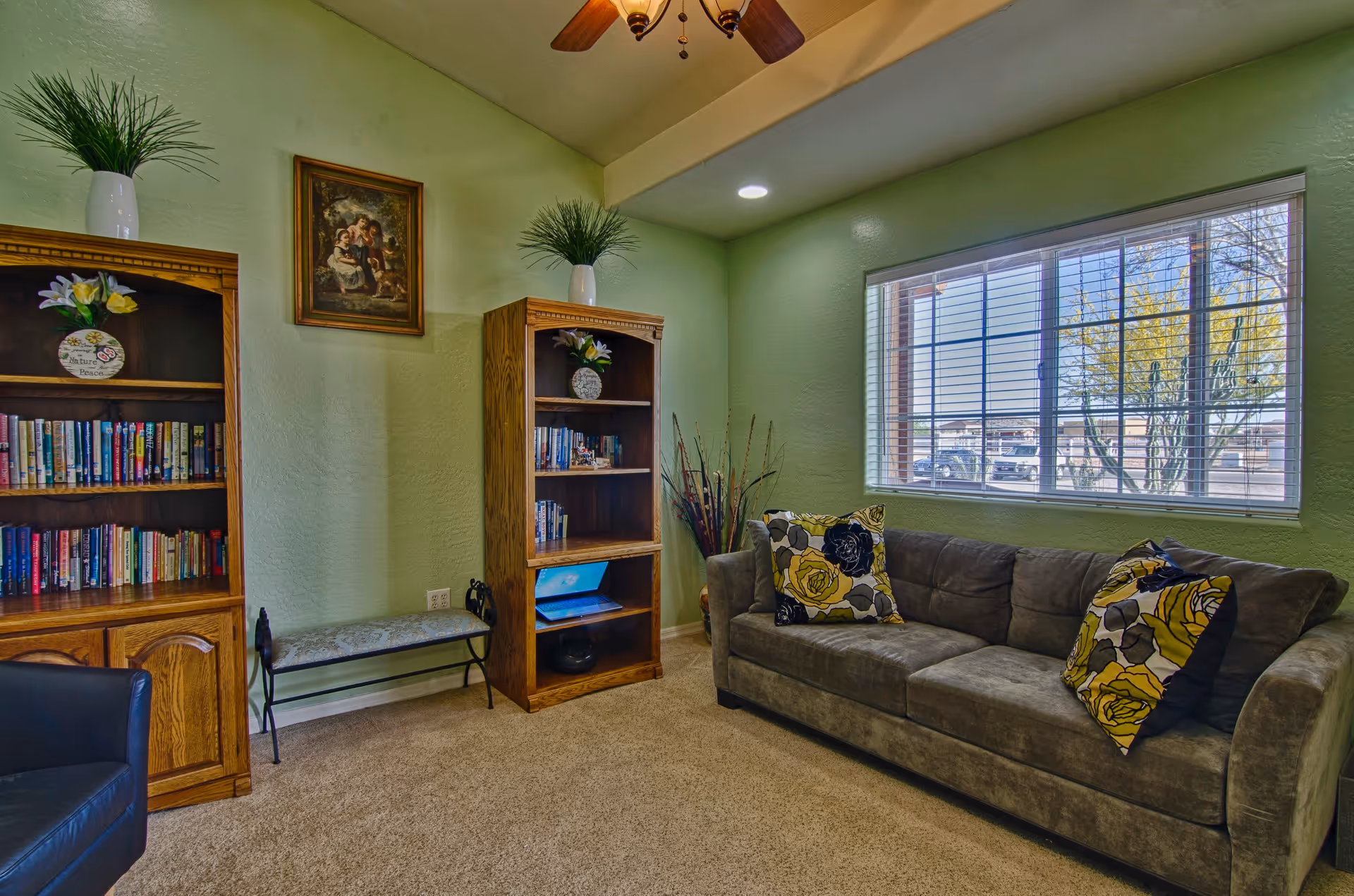 A cozy living room with a gray sofa adorned with floral patterned cushions, two wooden bookshelves filled with books and decorative items, a small bench, a ceiling fan with lights, and a large window with blinds letting in natural light. The walls are painted light green and the floor is carpeted.