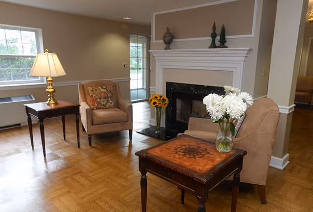 A cozy living room with two beige armchairs, a wooden side table with a lamp, and a wooden coffee table with a vase of white flowers. There is a fireplace with decorative items on the mantel and a vase of sunflowers on the hearth. Large windows and a glass door allow natural light into the room.