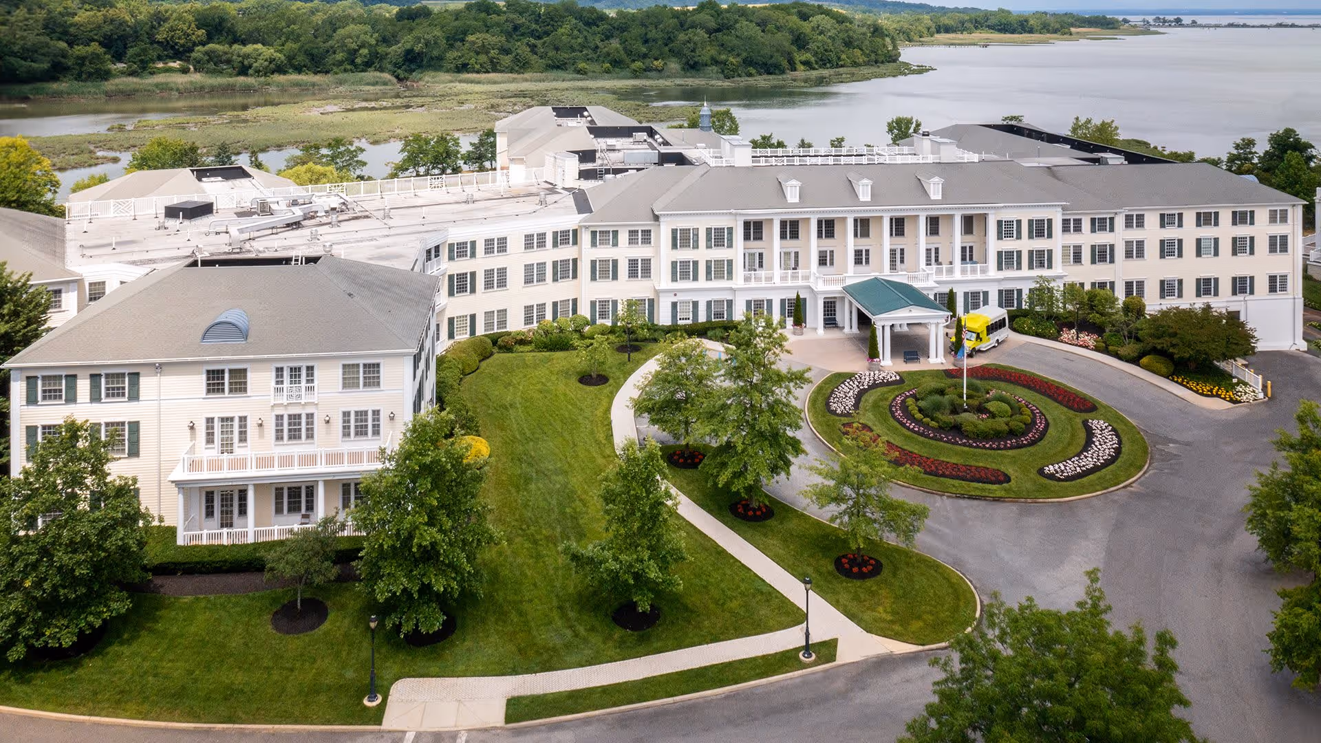 A large senior living facility building with white exterior walls and multiple windows, surrounded by well-maintained green lawns, trees, and landscaped flower beds. A circular driveway with a flagpole and a covered entrance is visible. The facility is located near a body of water with trees and greenery in the background.