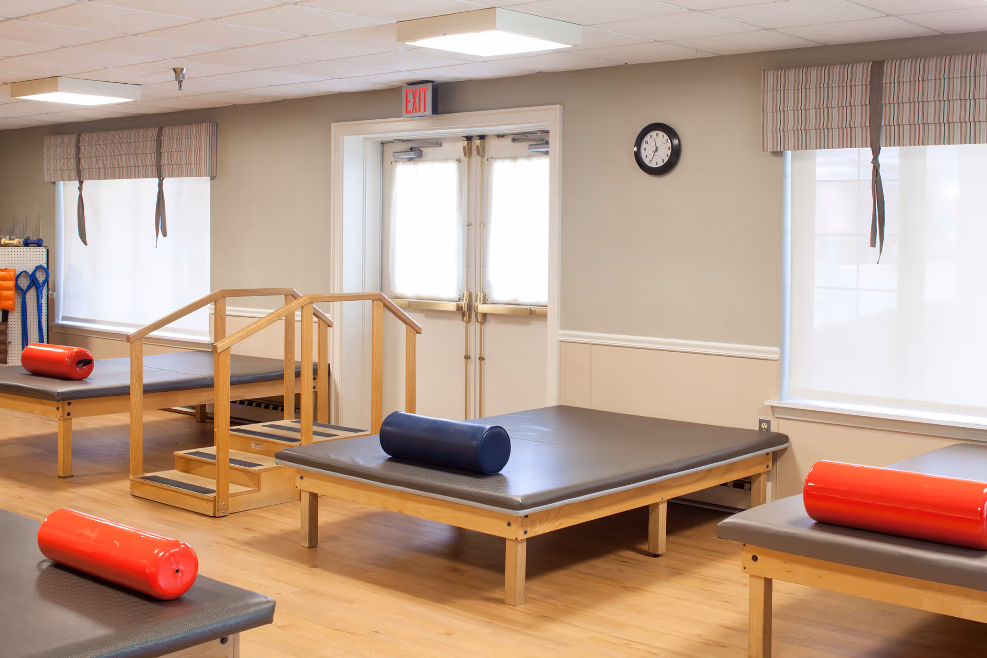A bright therapy room with padded wooden treatment tables, a set of practice stairs, and several red and blue foam rollers.