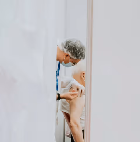 A healthcare worker wearing a hairnet, face mask, and gloves is administering a vaccine injection to an elderly person seated with their sleeve rolled up.