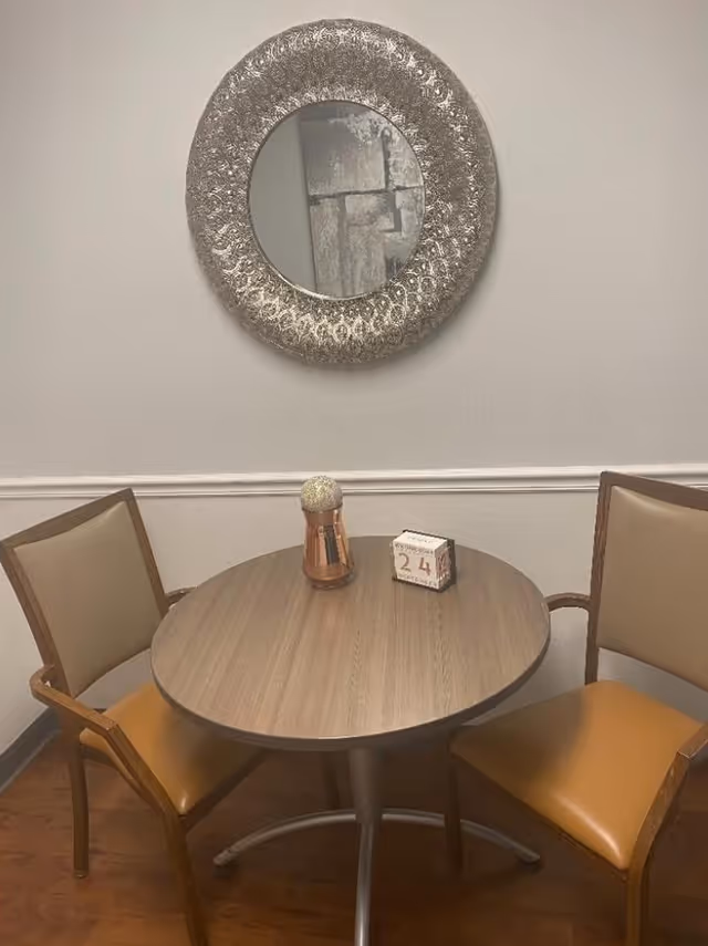 A small round wooden table with two tan cushioned chairs on either side. On the table, there is a decorative copper-colored vase with a textured spherical top and a small block calendar showing the date 24. Above the table, a large round ornate silver mirror hangs on a plain light-colored wall with a white chair rail.