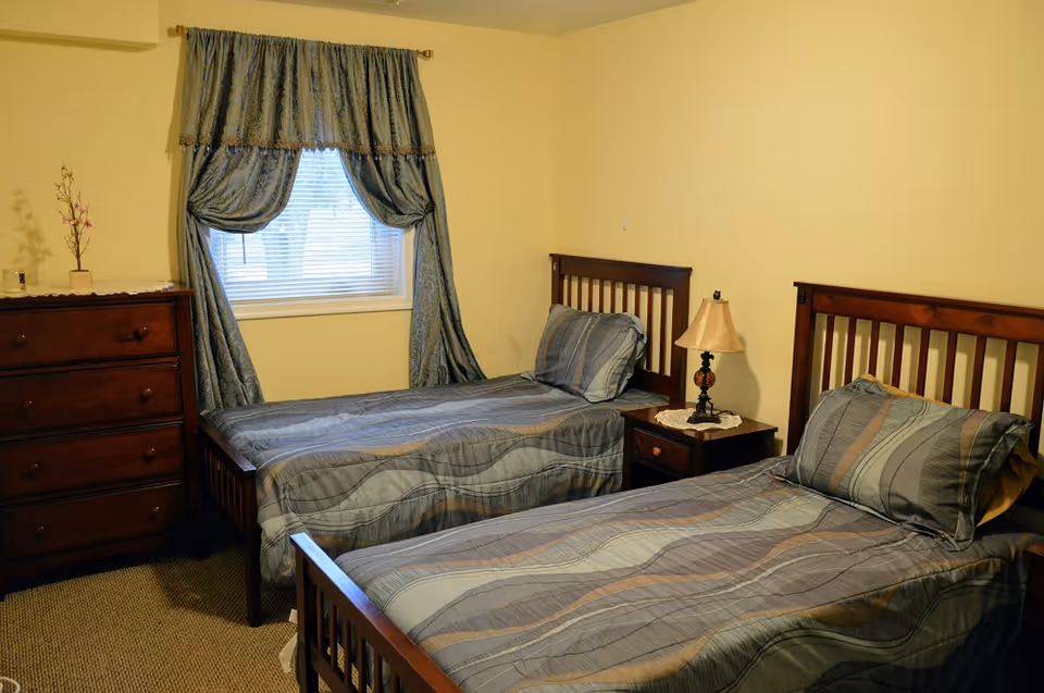 A bedroom with two twin beds featuring wooden headboards and matching striped bedding in shades of blue and gray. Between the beds is a wooden nightstand with a lamp on top. To the left of the beds is a wooden chest of drawers with a small decorative plant on it. A window with blue curtains is centered on the wall behind the beds.