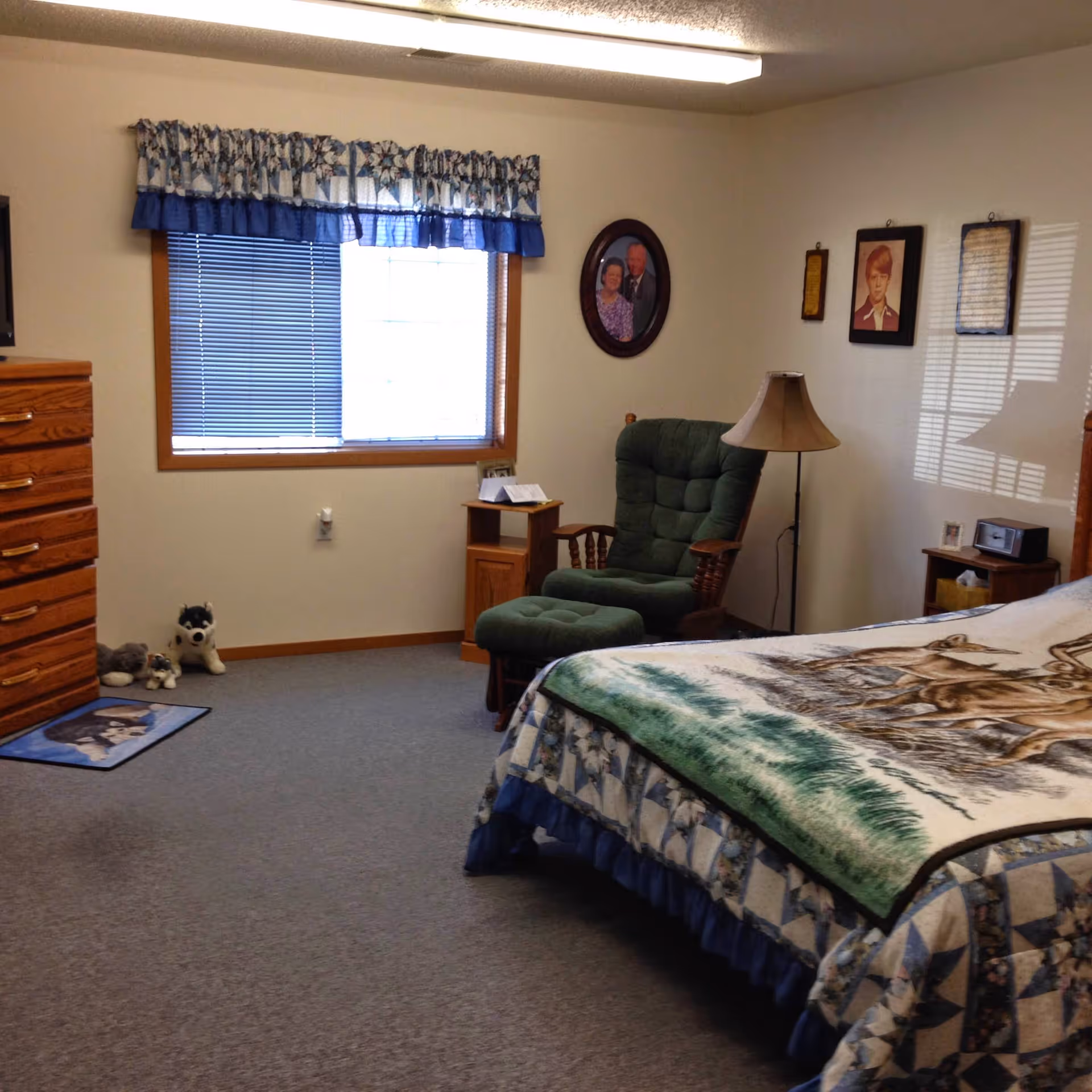 A tidy bedroom featuring a bed with patterned bedding, a green upholstered recliner and ottoman, wooden dressers and nightstands, a window with a blue valance, and framed photos on the wall.