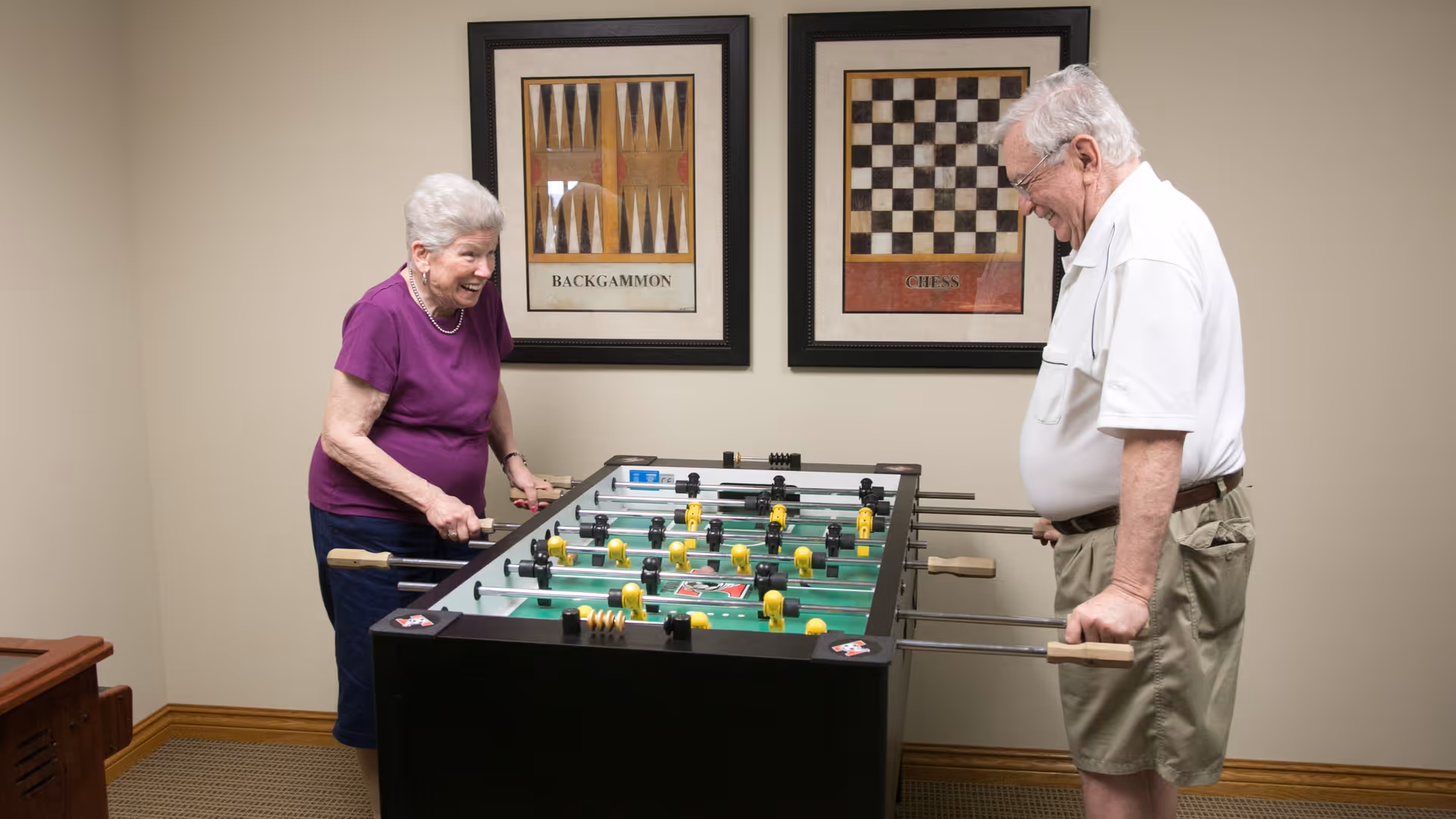 An elderly woman and an elderly man playing foosball in a room with beige walls. The woman is wearing a purple shirt and dark pants, and the man is wearing a white polo shirt and khaki shorts. Behind them on the wall are framed pictures of backgammon and chess boards.