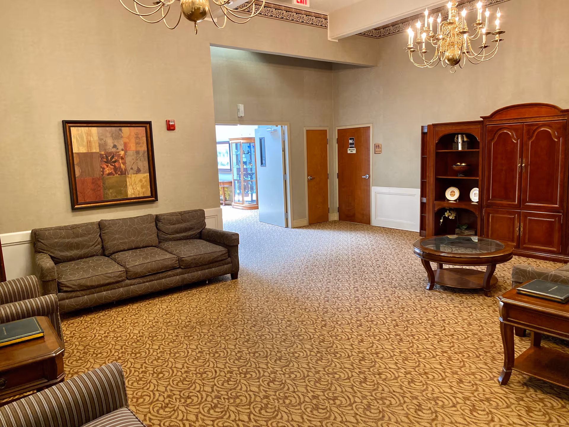 A spacious living room area in a senior living facility with beige patterned carpet, a brown sofa, two striped armchairs, a round glass-top coffee table, and a wooden cabinet with shelves displaying decorative plates. There is a framed abstract painting on the wall and two brass chandeliers hanging from the ceiling. Two closed wooden doors and an open doorway leading to a bright hallway are visible in the background.