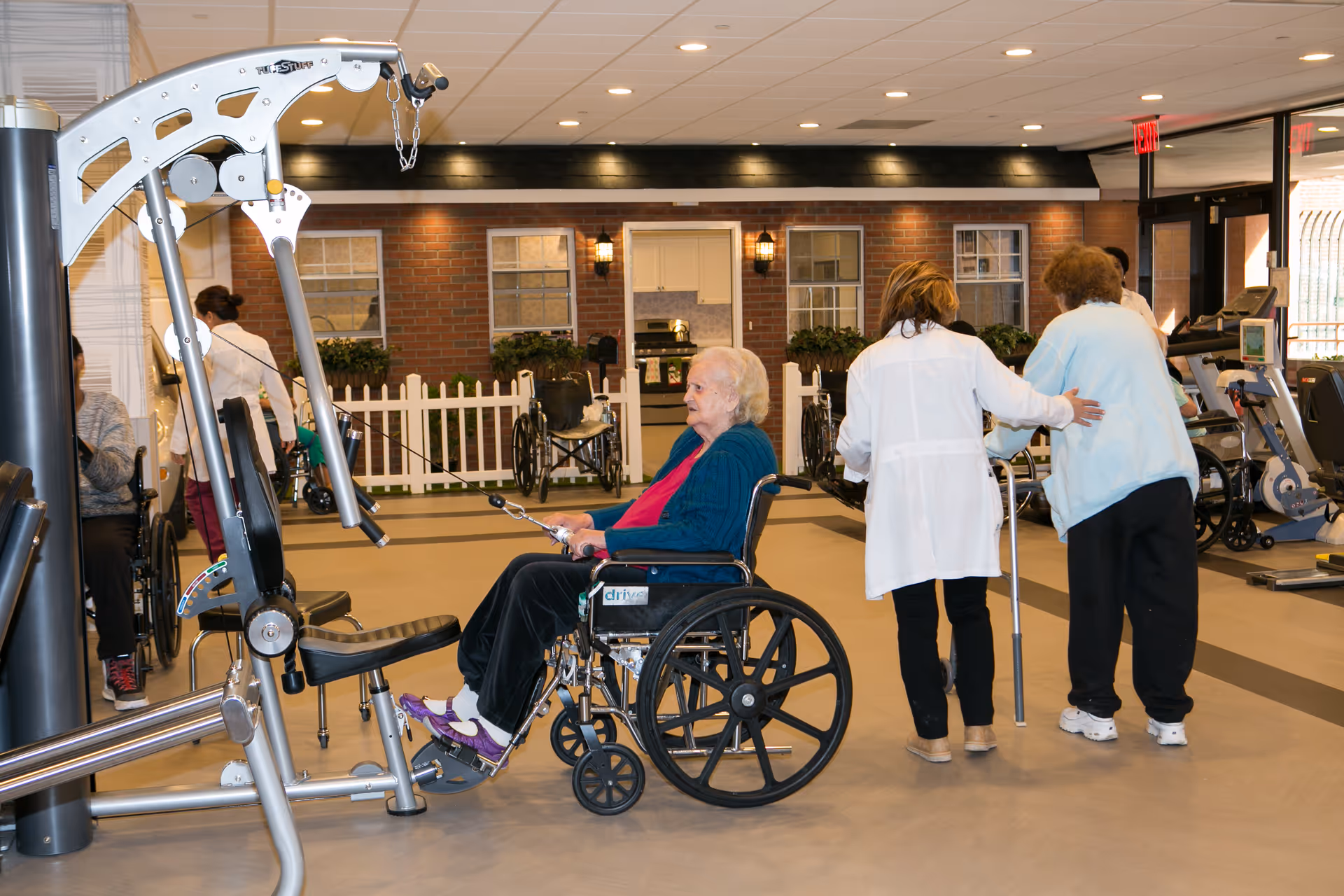An elderly woman in a wheelchair using exercise equipment in a rehabilitation gym area, with a caregiver assisting another elderly woman using a walker. The background shows a brick wall with windows and a white picket fence, and other exercise machines are visible.